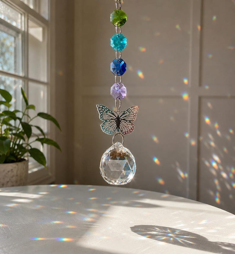 Op de afbeelding: Een hangende zonnevanger met een verticale ketting van vier gefacetteerde kralen in groen, turquoise, donkerblauw en lichtpaars. Onder de kralen hangt een zilverkleurige metalen vlinderbedel aan een helder, bolvormig gefacetteerd kristallen prisma.