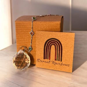 May include: A clear crystal prism hanging from a silver chain with colorful gemstones. The prism is in a brown box with a tag that says "Eternal Rainbows".