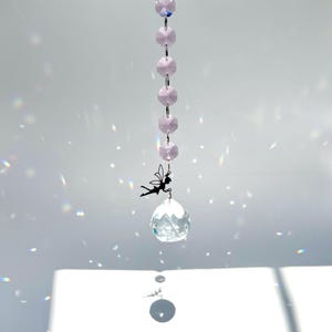 May include: A decorative crystal suncatcher with a faceted clear crystal ball and a small black metal fairy charm. The suncatcher is suspended from a chain of light pink beads.