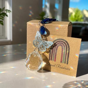 Op de afbeelding: Een kristallen zonnevanger met een zilverkleurige metalen vlinderbedel en donkerblauwe kralen, hangend voor een bruine kartonnen doos. Een bruine kaart met een regenboogafbeelding en de tekst Eternal Rainbows staat voor de doos.