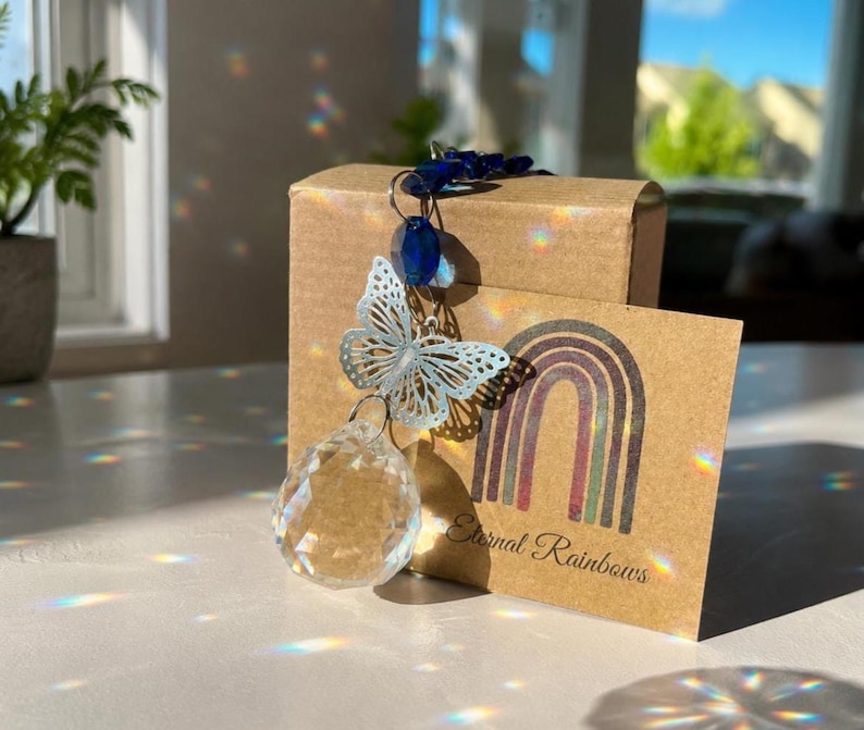 Op de afbeelding: Een kristallen zonnevanger met een zilverkleurige metalen vlinderbedel en donkerblauwe kralen, hangend voor een bruine kartonnen doos. Een bruine kaart met een regenboogafbeelding en de tekst Eternal Rainbows staat voor de doos.