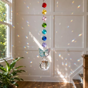 Op de afbeelding: Een hangende zonnevanger met een verticale streng van zeven gefacetteerde, veelkleurige kralen in rood, geel, roze, groen, turquoise, blauw en paars. Onder de kralen hangt een zilverkleurige vlinderbedel boven een helder, bolvormig kristal.