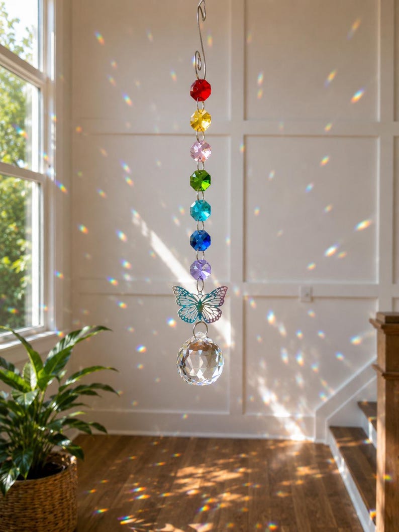 Op de afbeelding: Een hangende zonnevanger met een verticale streng van zeven gefacetteerde, veelkleurige kralen in rood, geel, roze, groen, turquoise, blauw en paars. Onder de kralen hangt een zilverkleurige vlinderbedel boven een helder, bolvormig kristal.