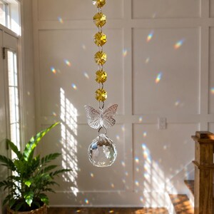 Op de afbeelding: Een hangende zonnevanger met een verticale streng van zes gefacetteerde gele kristallen kralen, een doorzichtige vlindervormige bedel en een grote heldere bolvormige kristallen prisma die regenboogkleurige lichtreflecties werpt.