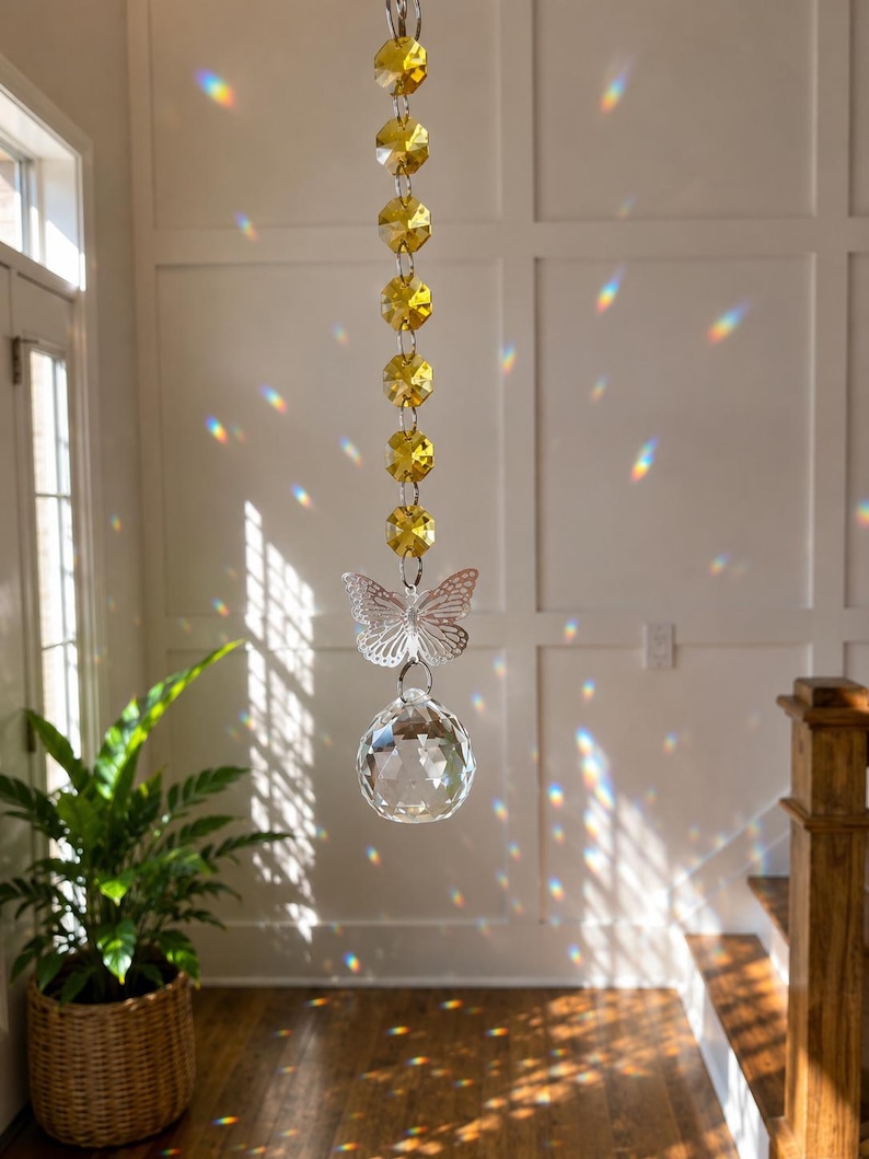 Op de afbeelding: Een hangende zonnevanger met een verticale streng van zes gefacetteerde gele kristallen kralen, een doorzichtige vlindervormige bedel en een grote heldere bolvormige kristallen prisma die regenboogkleurige lichtreflecties werpt.