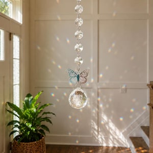 Op de afbeelding: Een hangende zonnevanger met een verticale ketting van heldere, gefacetteerde glaskralen, een zilverkleurige metalen vlinderbedel met gedetailleerde uitgesneden vleugels en een grote, heldere, bolvormige kristallen prisma onderaan.