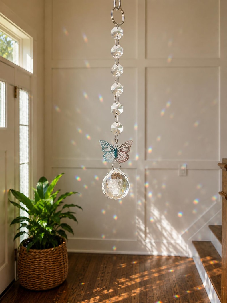 Op de afbeelding: Een hangende zonnevanger met een verticale ketting van heldere, gefacetteerde glaskralen, een zilverkleurige metalen vlinderbedel met gedetailleerde uitgesneden vleugels en een grote, heldere, bolvormige kristallen prisma onderaan.