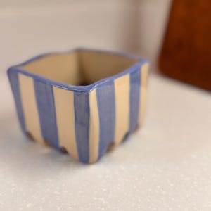 May include: A square ceramic planter with a light beige interior and exterior. The exterior features vertical blue stripes. The planter is sitting on a white speckled surface. The planter is approximately 4 inches wide.