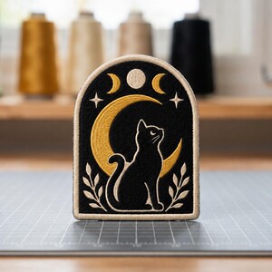 May include: An arched embroidered patch featuring a black cat sitting in front of a large gold crescent moon. Above the cat are two smaller gold crescent moons and a central circle. The design includes two white leafy branches and four-pointed stars.