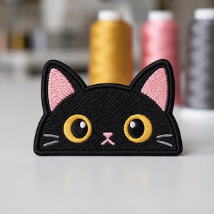 May include: An embroidered patch shaped like a black cat head with large yellow eyes, pink inner ears, a small pink nose, and grey whiskers. The patch features a thick black border and is designed with a flat bottom edge for placement on fabric.