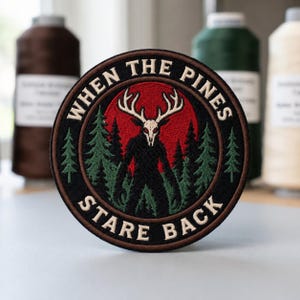 May include: A circular embroidered patch featuring a black silhouette of a creature with a white deer skull head standing in a forest of green pine trees against a red circular background, with the text "WHEN THE PINES STARE BACK" in white.
