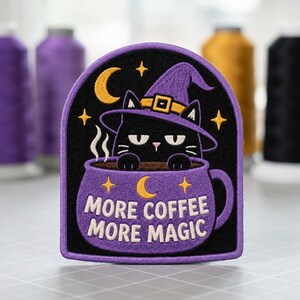 May include: An embroidered patch with a black background and a purple border. It features a black cat wearing a purple witch hat, sitting inside a purple coffee mug. The text reads MORE COFFEE MORE MAGIC, accompanied by yellow stars and a crescent moon.