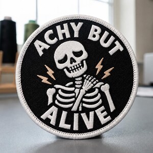 May include: A circular embroidered patch with a black background and a white border. The design features a white skeleton holding its shoulder with two tan lightning bolt icons. The text reads ACHY BUT ALIVE in bold white capital letters.