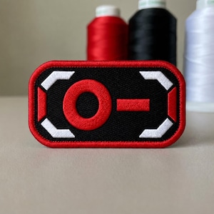 May include: A rectangular embroidered patch with rounded corners featuring a black background, a red circular emblem, a red horizontal dash, and white geometric corner accents, all bordered by a thick red stitched edge.