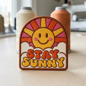 May include: An embroidered patch with an arched top featuring a yellow smiling sun with pink cheeks, surrounded by orange and pink rays. Below the sun are white clouds and the text STAY SUNNY in orange and yellow bubble letters with a brown border.
