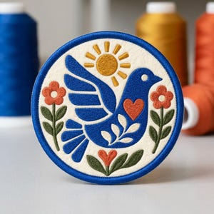 May include: A circular embroidered patch featuring a blue bird with a red heart on its chest, surrounded by orange flowers with green stems and a yellow sun, all set against an off-white background with a thick blue border trim.