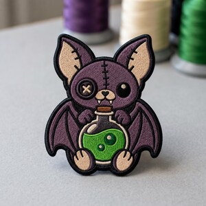 May include: An embroidered patch featuring a purple bat with large ears, a button eye, and a stitched forehead. The bat holds a round glass bottle filled with bright green liquid, featuring a brown cork stopper and two small white fangs visible.