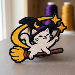 May include: An embroidered patch featuring a white cat wearing a black witch hat with a yellow crescent moon. The cat is riding a brown broomstick with yellow bristles and holding a yellow star-tipped wand, with a yellow swoosh design.