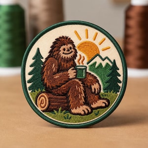 May include: A circular embroidered patch featuring a brown Bigfoot sitting on a log, holding a green mug with steam rising. The scene includes a yellow sun, green pine trees, and white mountain peaks against a cream-colored background.