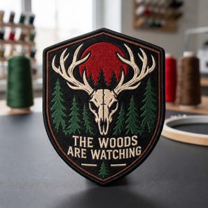 May include: A shield-shaped embroidered patch featuring a beige deer skull with antlers, a red sun, and green pine trees on a black background. The text "THE WOODS ARE WATCHING" is stitched in beige at the bottom with a small green pine tree icon.