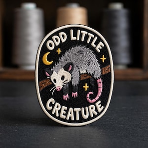 May include: An oval embroidered patch with a black background featuring a gray opossum hanging from a brown branch. The text ODD LITTLE CREATURE is stitched in white letters. A yellow crescent moon and small stars surround the opossum.