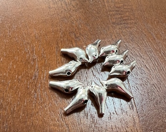 May include: A group of silver bird-shaped charms arranged in a circle on a wooden surface. The charms have a smooth, metallic finish and a small hole at the top for attaching to jewelry or other crafts.