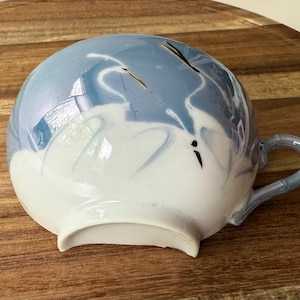 Op de afbeelding: Een keramische theekop met een lichtblauw en wit ontwerp. De kop heeft een gebogen handvat en is versierd met een geschilderde scène van witte vogels tegen een blauwe lucht. De kop staat op een houten oppervlak.