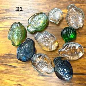 May include: A collection of ten glass sea life decorations, including fish and seashells. The decorations are in various shades of green, blue, and clear. The decorations are arranged on a wooden surface.