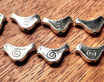 May include: Ten silver bird-shaped beads with a swirl design. The beads are about 1 inch long and have a slightly textured surface.