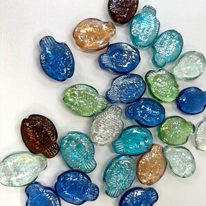May include: A collection of small, flat, fish-shaped glass beads in various translucent colors including blue, green, brown, and clear. Each bead features a molded scale pattern on the surface and a small hole at the top for threading or crafting.