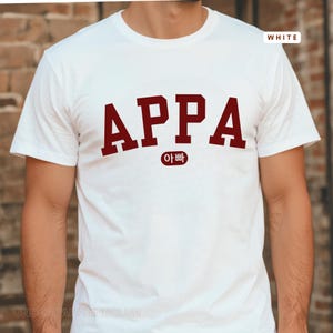 May include: White t-shirt with the word "APPA" in maroon block letters, with the Korean word "아빠" underneath. The word "WHITE" is in the upper right corner. The shirt is a crew neck.