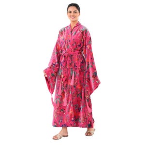 Puede incluir: Una bata rosa brillante con estampado floral, mangas largas y cinturón a juego. La bata presenta un patrón botánico con acentos verdes, azules y rojos. Borlas cuelgan de las mangas y el cinturón. La bata está hecha de una tela suave y fluida.