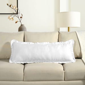 May include: A white rectangular pillow with a ruffled edge, resting on a beige sofa. The pillow has the words "Cotton Print Club" embroidered on it. A lamp and a vase with flowers are in the background.