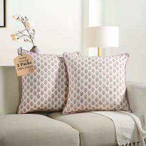 Op de afbeelding: Twee decoratieve sierkussens met een witte achtergrond en een herhalend patroon van roze en bruine bloemmotieven. De kussens hebben een roze rand en liggen op een beige bank. Een label luidt "Cotton Print Club 2 Packs 100% Cotton."