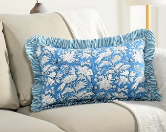 May include: A rectangular blue pillow with a white floral and bird pattern. The pillow has a ruffled edge with blue and white stripes. The pillow is on a beige sofa.