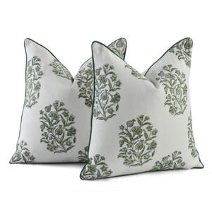 Könnte beinhalten: Zwei weiße Zierkissen mit einem grünen Blumenmuster. Die Kissen haben eine dunkelgrüne Paspel an den Rändern. Das florale Design ist ein sich wiederholendes Muster aus stilisierten Blumen und Blättern.