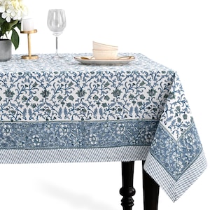 May include: A white and blue floral tablecloth with a border of blue and white stripes. The tablecloth is on a black table with a white background. A wine glass, plates, and flowers are on the table.
