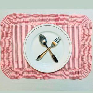 Peut inclure: Une assiette blanche avec un bord doré, une cuillère et une fourchette croisées, posées sur un set de table à rayures rouges et blanches avec un bord à volants. Le set de table est rectangulaire.