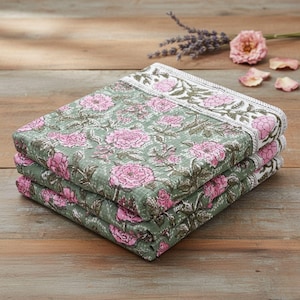 May include: A stack of three folded floral patterned blankets. The blankets feature a repeating pattern of pink flowers and green leaves on a light green background. The edges have a white border with pink floral designs. The blankets are stacked on a wooden surface.