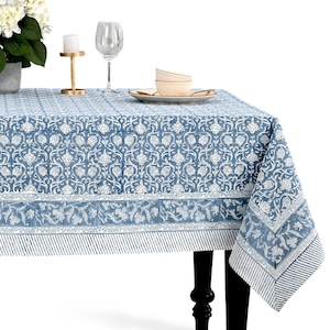 Puede incluir: Un mantel azul y blanco con estampado floral cubre una mesa. El mantel tiene un diseño detallado con un borde de rayas y motivos florales. Un candelabro dorado, una copa de vino y un plato con cuencos están sobre la mesa.