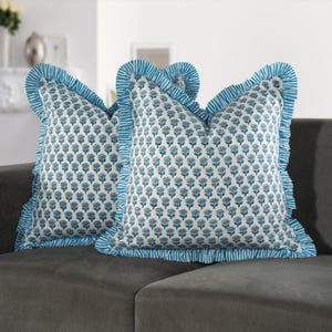 Puede incluir: Dos almohadas de tiro azul y blanco con estampado floral y ribete con volantes. Las almohadas están en un sofá gris.