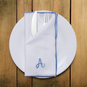 May include: A white linen napkin with a blue embroidered border and the letter 'A' in blue thread. The napkin is folded in half and placed on a white plate with a silver knife and fork inside.