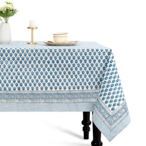 May include: A rectangular tablecloth featuring a repeating blue floral block print pattern on a white background, bordered by a decorative floral band and a light blue and white striped edge, draped over a table with dark wooden legs.