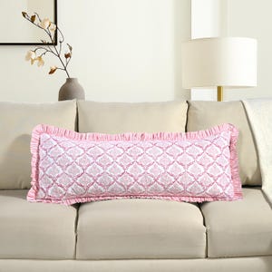 Puede incluir: Un cojín decorativo rectangular con un estampado de damasco blanco y rosa. El cojín tiene un borde rosa con volantes. El cojín está colocado sobre un sofá beige. Un jarrón con un arreglo floral está en el fondo.