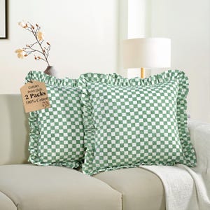 Op de afbeelding: Twee vierkante kussens met een groen-wit geblokt patroon en gerimpelde randen. De kussens liggen op een beige bank. Een label leest "Cotton Print Club 2 Packs 100% Cotton."