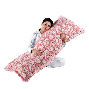 Pode incluir: Uma almofada de corpo longa e retangular na cor coral com um padrão floral e de pássaros brancos. A almofada tem uma borda com babados. A pessoa que segura a almofada está vestindo uma camisa e calças brancas. A almofada foi projetada para conforto e suporte.