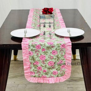 May include: A floral table runner with a light green background and pink rose pattern. The runner has a ruffled pink border. Two white plates with forks and knives are set on the table. A mirrored box with red flowers sits in the center.