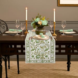 Puede incluir: Una mesa de comedor preparada para una comida, con un camino de mesa con estampado floral verde y blanco. La mesa está puesta con platos, cubiertos, copas de vino y un jarrón con flores. Dos velas encendidas en candelabros dorados.