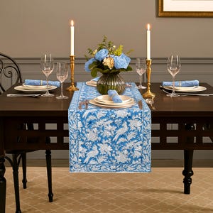 Peut inclure: Une table à manger dressée avec un chemin de table bleu et blanc à motifs floraux. La table est ornée d'assiettes, de couverts et de verres à vin. Deux bougies allumées dans des bougeoirs dorés encadrent un vase de fleurs bleues.