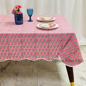 Puede incluir: Un mantel rosa floral con borde festoneado, sobre una mesa de madera. El mantel presenta un patrón repetitivo de diseños florales verdes y blancos. Un jarrón de cristal azul con flores rosas, dos platos blancos y un vaso azul están sobre la mesa.